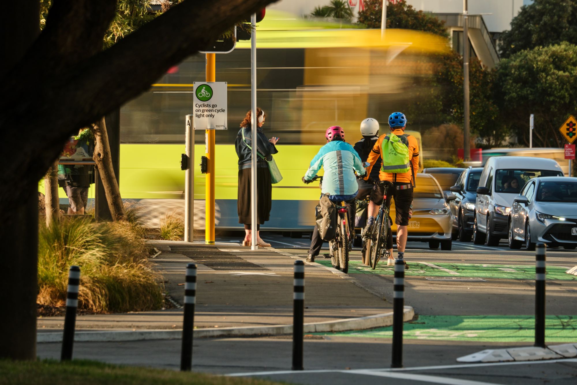 3 cyclists wait at a crossing for a moving bus to pass.